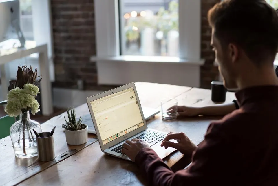 A man working on a laptop at a wooden desk by a window