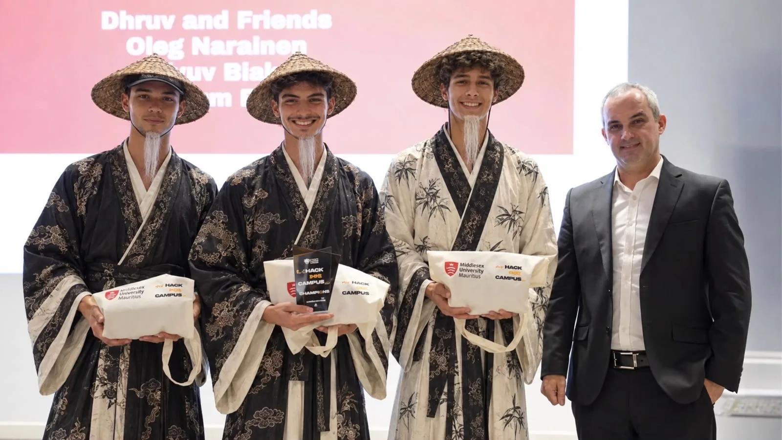 Three Black Reef founders in matching robes and straw hats holding the Hack the Campus champions trophy, next to a member of the judging panel.